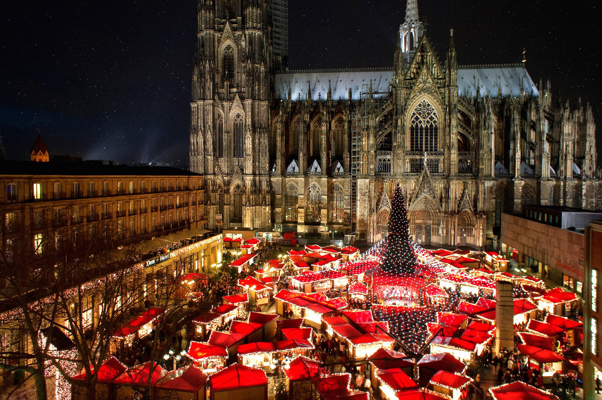 An enchanting aerial view of the Cologne Cathedral Christmas Market in Germany, set against the backdrop of Cologne's iconic Gothic cathedral at night, with twinkling lights creating a magical atmosphere. Perfect for luxury Christmas travel and private jet charters to European Christmas markets.