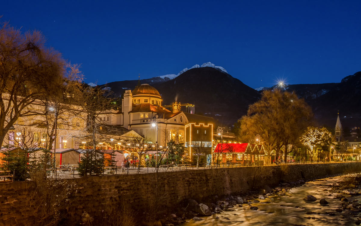 A scenic view of the Merano Christmas Market in Italy, with festive stalls and decorations set against the stunning backdrop of snow-capped mountains, perfect for luxury Christmas travel to European Christmas markets.