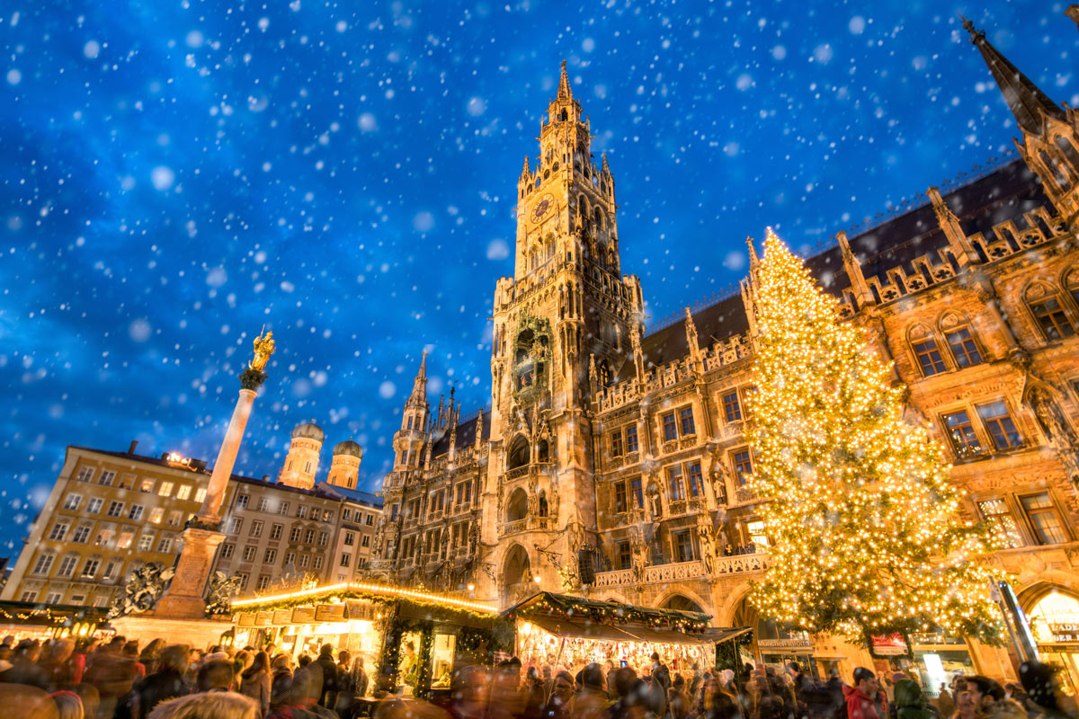 A picturesque view of the Munich Christkindlmarkt at Marienplatz, with soft snowflakes falling and visitors browsing the festive Bavarian Christmas market, a perfect destination for luxury Christmas travel to European Christmas markets.